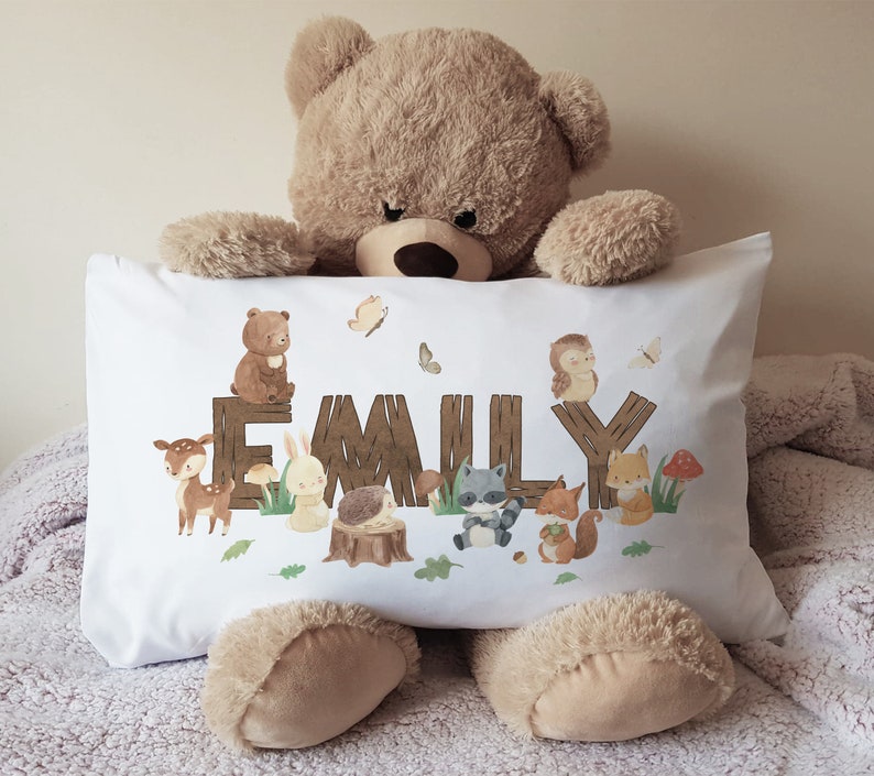 May include: White pillowcase with a woodland animal theme. The pillowcase features a brown bear, a deer, a rabbit, a raccoon, a hedgehog, and a fox. The name "EMILY" is spelled out in brown letters on a wooden fence.