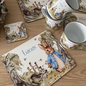 May include: Four personalized placemats with a Peter Rabbit theme. Each placemat features a different name, "Olivia", "Lucas", "Lucas", and "Maddie". The placemats are white with a watercolor illustration of Peter Rabbit in a garden setting. The placemats are paired with four enamel mugs, each with a different name.