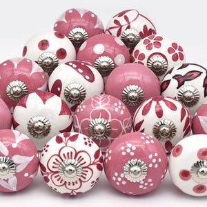 Könnte beinhalten: Eine Sammlung dekorativer Keramik-Schrankknöpfe in verschiedenen Rosa- und Weißtönen. Die Knöpfe weisen Blumen-, Tupfen- und Streifenmuster mit silberfarbenen Metallbeschlägen auf. Die Knöpfe sind vor weißem Hintergrund pyramidenförmig angeordnet.