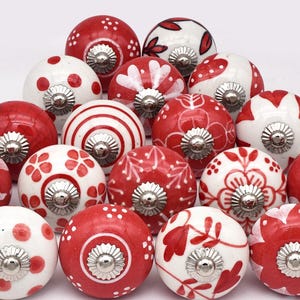 Peut inclure: Ensemble de boutons de meubles en céramique rouge et blanc avec divers motifs, dont des pois, des rayures et des motifs floraux. Chaque bouton a une base en métal argenté. Les boutons sont disposés sur fond blanc.