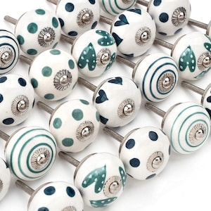 Könnte beinhalten: Eine Sammlung dekorativer Keramik-Schrankknöpfe. Die Knöpfe sind weiß mit verschiedenen Mustern in Blau und Grün, darunter Punkte, Streifen und Blattmotive. Jeder Knopf hat einen silbernen Metallsockel.