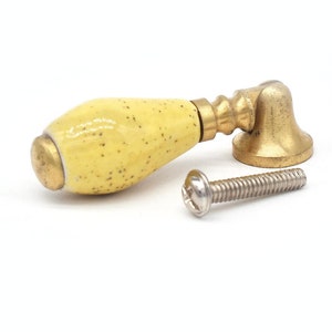 Op de afbeelding: Een gele keramische kastknop met een gouden metalen basis en een schroef.