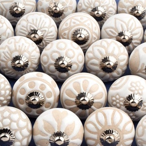 May include: A collection of 25 white ceramic cabinet knobs with various embossed floral and geometric designs. The knobs have a silver-toned metal base.