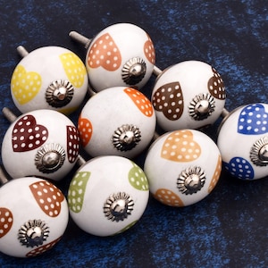 Pode incluir: Uma coleção de dez botões de armário de cerâmica com base branca e corações de bolinhas de várias cores. Os botões têm um parafuso prateado no centro.