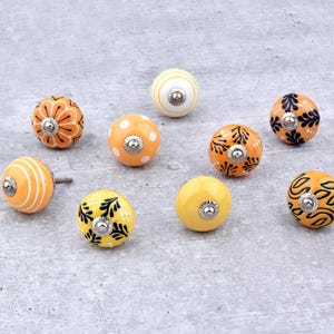 Könnte beinhalten: Eine Sammlung dekorativer Keramik-Schrankknöpfe in verschiedenen Farben und Mustern. Die Knöpfe weisen Designs wie Streifen, Tupfen und florale Motive in Orange-, Gelb- und Weißtönen auf. Jeder Knopf hat eine silberne Metallbasis.