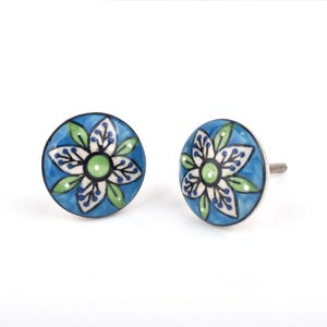 Op de afbeelding: Twee blauwe keramische kastknoppen met een bloemmotief. De knoppen hebben een witte bloem met groene en zwarte accenten, tegen een blauwe achtergrond. De knoppen hebben een zilveren schroef voor installatie.