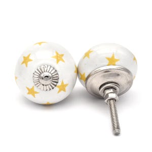 May include: Two white ceramic cabinet knobs with a yellow star pattern. The knobs have a silver metal base and screw.