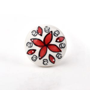 May include: A round ceramic knob with a white base featuring a central red four-petaled flower design. The surface is decorated with smaller red leaf-shaped accents and black circular patterns surrounding the central floral motif.