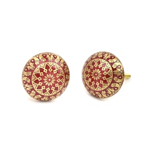 May include: Two decorative cabinet knobs. Each knob is round with a red centre and gold-coloured filigree design. The knobs have a brass-coloured stem for mounting. The knobs are likely made of metal or ceramic.