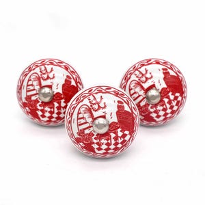May include: Three white ceramic cabinet knobs with a red design featuring a checkered pattern and a decorative scene. Each knob has a silver-coloured metal screw. The knobs are round and have a decorative edge.