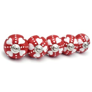 May include: Five red ceramic cabinet knobs with white heart and dot designs. Each knob has a silver metal base.