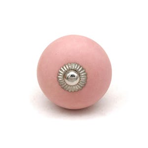 May include: A pale pink ceramic cabinet knob with a silver-coloured, flower-shaped metal accent in the centre. The knob is set against a white background, highlighting its simple design and colour.