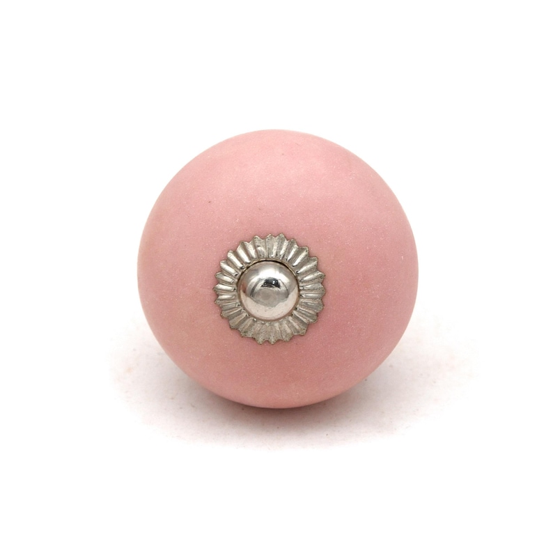 May include: A round, light pink ceramic cabinet knob with a silver-colored, flower-shaped metal accent in the center. The knob is set against a white background, highlighting its simple design and color.