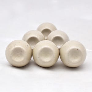 Op de afbeelding: Zes crèmekleurige keramische knoppen gerangschikt in een piramidevorm. Elke knop heeft een afgeronde bovenkant en een verzonken basis. De knoppen zijn glad en hebben een matte afwerking, geschikt voor meubels of decoratieve projecten.