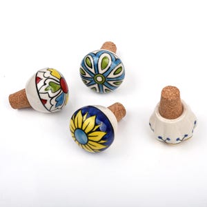 May include: Four decorative bottle stoppers with cork bases. The stoppers feature hand-painted ceramic designs in various colors, including blue, yellow, red, and green. The designs include floral and geometric patterns.