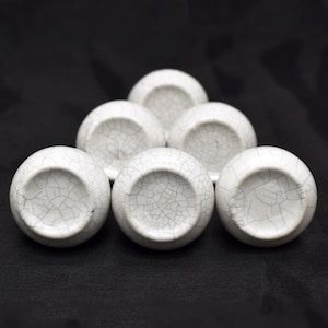Peut inclure: Six boutons de porte en céramique blanche avec un motif de glaçure craquelée, disposés en triangle sur fond noir. Les boutons sont ronds avec une base plate et présentent des motifs de fissures uniques.
