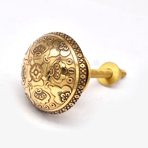 Może przedstawiać: Złoty uchwyt do szafki z ozdobnym kwiatowym wzorem. Gałka jest okrągła ze szczegółowym wzorem kwiatów i liści. Krawędzie mają powtarzający się wzór kół. Śruba jest również złota.