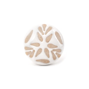 Op de afbeelding: Een ronde keramische meubelknop met een glanzende witte glazuurafwerking en een herhalend patroon van onbedekte, beige, druppelvormige kleivormen die in een radiaal ontwerp over het oppervlak van het ronde beslag zijn gerangschikt.