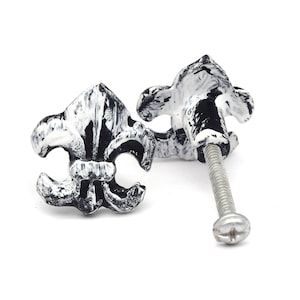 Puede incluir: Dos tiradores de armario con forma de flor de lis con un acabado en blanco y negro envejecido. Los tiradores presentan un diseño decorativo e incluyen un tornillo plateado para la instalación. Ideales para añadir un toque de elegancia a los muebles.