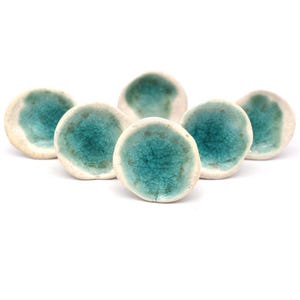 May include: Six ceramic knobs with a white base and a teal blue glaze. The knobs are round and have a slightly irregular shape.