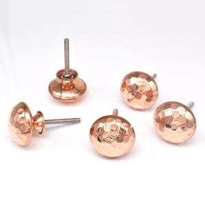 Może przedstawiać: Pięć miedzianych gałek do szafek z młotkowaną fakturą. Każda gałka ma okrągły, wypukły kształt i gwintowany trzpień ułatwiający montaż. Gałki mają ciepły, metaliczny kolor, idealny do dodania elegancji każdemu meblowi.