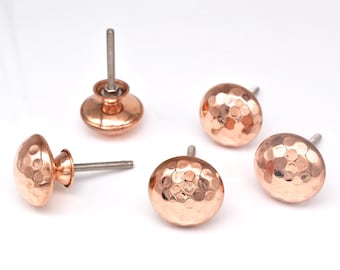 May include: Five copper-colored cabinet knobs with a hammered texture. Each knob has a round, domed shape and a threaded post for easy installation. The knobs are a warm, metallic color, perfect for adding a touch of elegance to any furniture piece.