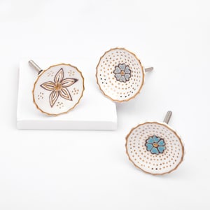 Op de afbeelding: Drie ronde keramische ladeknoppen met geschulpte randen en goudkleurige afwerking. Eén knop heeft een centraal bruin bloemmotief, terwijl de andere twee kleine blauwe bloemen tonen, omringd door een patroon van kleine gouden stippen op wit.