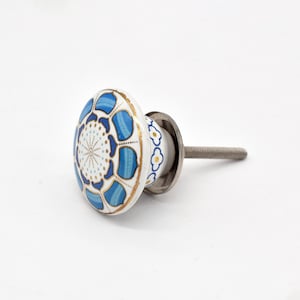 Puede incluir: Un pomo de cerámica redondo para muebles con base blanca y un patrón floral en azul y dorado en la cara frontal. Los laterales del pomo están decorados con un motivo repetitivo de flores azules y amarillas, e incluye un perno de metal plateado.