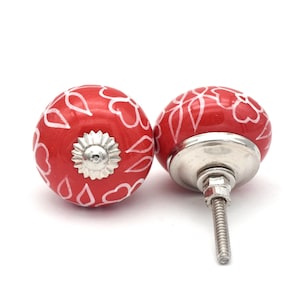Op de afbeelding: Twee rode keramische kastknoppen met witte bloemmotieven. De knoppen hebben een zilveren metalen basis en een schroef.