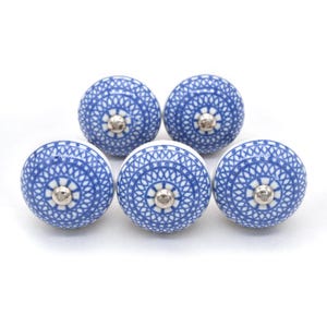May include: Five round ceramic cabinet knobs. Each knob is blue and white with a detailed, symmetrical pattern. The knobs have a silver-colored metal base. The knobs are likely used for furniture or cabinet doors.