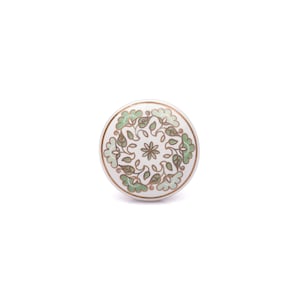 Può includere: Un pomello rotondo in ceramica per mobili con base bianca caratterizzato da un motivo floreale centrale circondato da un disegno circolare di foglie e petali verde chiaro e marroni, delineato da sottili linee marroni sulla superficie.