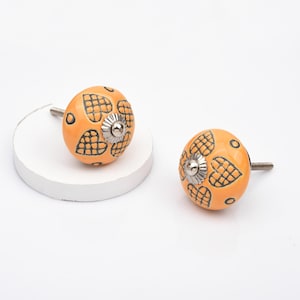 May include: Two round orange cabinet knobs featuring a black grid-patterned heart design. Each knob has a silver-toned metal centre accent and a threaded metal screw extending from the back for installation on furniture or cabinetry doors.