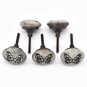 May include: Five decorative cabinet knobs. The knobs have a distressed, crackled white finish with a black butterfly design. The hardware is dark metal. The knobs are for furniture and home decor.