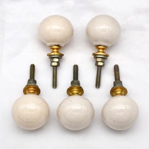 Può includere: Cinque pomelli per mobili in ceramica bianco sporco con ferramenta color ottone. I pomelli sono sferici e hanno una vite per il fissaggio. L'hardware include una base color ottone e un dado per fissare il pomello.