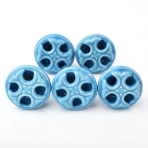 May include: Five light blue ceramic cabinet knobs with a decorative, openwork design. Each knob features a circular shape with a glossy finish and a pattern of curved cutouts. The knobs are arranged on a white background.