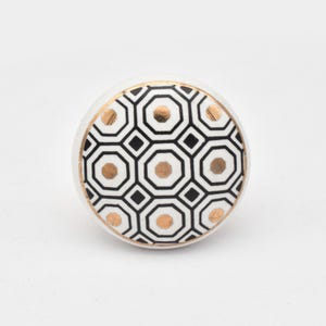 May include: White ceramic cabinet knob with a geometric pattern in black and gold. The knob features a circular design with octagons and squares, accented by a gold border. This decorative hardware adds a touch of modern style to furniture.