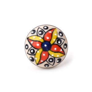 Puede incluir: Un pomo de cerámica redondo con un diseño floral pintado a mano que presenta un punto azul oscuro central rodeado de pétalos rojos y amarillos alternos. El fondo de color crema está decorado con pequeños círculos negros y puntos blancos.