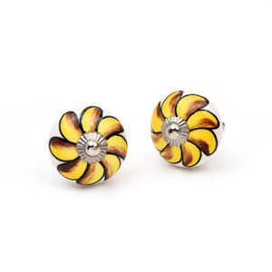May include: Two round ceramic cabinet knobs featuring a hand-painted floral pattern with yellow and brown petals radiating from a central silver-toned metal hardware piece. The knobs have a white base and a smooth, glossy finish on the surface.