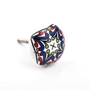 Può includere: Pomello per armadio in ceramica bianca con un motivo floreale decorativo rosso, blu e verde. Il pomello ha una vite argentata per il fissaggio. Il design è simmetrico e centrato sulla faccia del pomello.