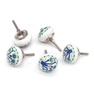 Puede incluir: Cinco pomos de cerámica blanca con un diseño floral en azul y verde. Cada pomo tiene una base y un tornillo de metal plateado. Los pomos están dispuestos sobre un fondo blanco.