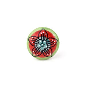 May include: A round ceramic knob featuring a hand-painted floral design. The center displays a light blue flower with black outlines, surrounded by a vibrant red circular band and a light green outer border with small white dot accents.