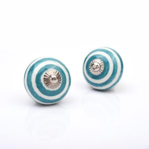 Puede incluir: Dos pomos de armario redondos de cerámica con un patrón de círculos concéntricos en color verde azulado y blanco brillante. Cada pomo está centrado con una tapa de metal plateado pulido con un borde festoneado y un pequeño detalle de cúpula.