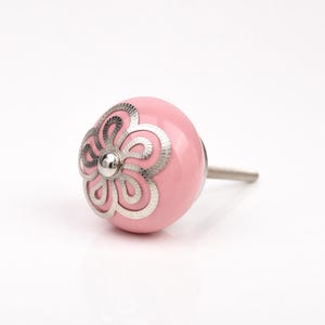 Könnte beinhalten: Ein rosa Keramik-Schrankknopf mit silbernem Blumendesign. Der Knopf hat eine runde Form und einen Metallstift zur Befestigung. Das florale Design ist detailliert und verleiht Möbeln eine dekorative Note.