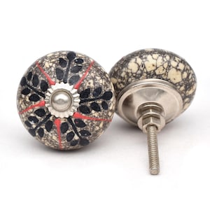 Puede incluir: Dos tiradores decorativos de cerámica para armarios. Los tiradores son redondos con un fondo moteado gris y crema. Cada tirador presenta un diseño de hojas negras con detalles rojos y una base de metal plateado. Para herrajes de muebles.