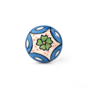 May include: A round ceramic knob featuring a central green four-petaled flower design on a cream background. The pattern is surrounded by blue curved shapes and small orange dots, with black outlines defining the floral and geometric motifs.