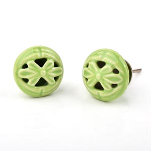 May include: Two light green ceramic cabinet knobs with a floral design. Each knob has a round shape with a flower-shaped cutout in the center. The knobs have a shiny finish and are mounted on metal hardware.