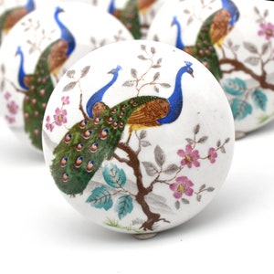 May include: White ceramic knob with a painted design of two peacocks with blue, green, and gold feathers on a branch with pink flowers.