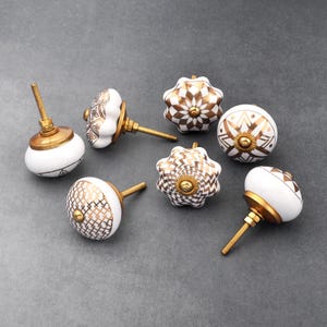 May include: Eight white ceramic cabinet knobs with gold geometric patterns. The knobs have a brass screw for attaching to a cabinet door.