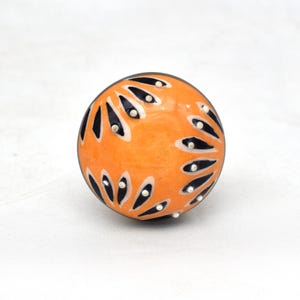 May include: Orange ceramic knob with a black and white geometric design. The knob has a glossy finish and is decorated with small white dots.