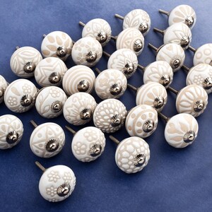 collection of white ceramic drawer knobs with intricate floral patterns and silver accents. The knobs are arranged in a basket lined with peach-colored fabric.